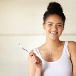 Dark-haired woman smiles while holding her toothbrush as she ponders how to improve our oral hygiene