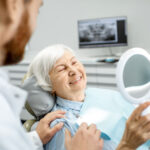 senior woman sitting in the dentist chair holds a mirror to look at her smile as the dentist looks on