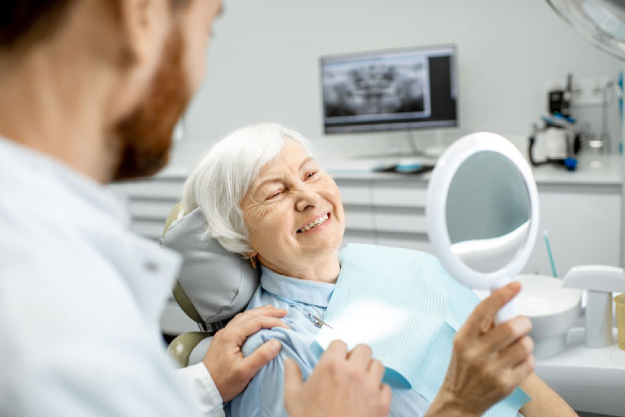 senior woman sitting in the dentist chair holds a mirror to look at her smile as the dentist looks on